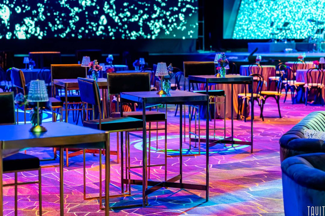 A modern event space with high tables and chairs, dim lighting, and vibrant blue and purple hues reflecting on the floor—perfect for Event Photography. Small flower arrangements and lamps adorn each table, with a large screen displaying abstract patterns.