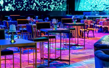 A modern event space with high tables and chairs, dim lighting, and vibrant blue and purple hues reflecting on the floor—perfect for Event Photography. Small flower arrangements and lamps adorn each table, with a large screen displaying abstract patterns.