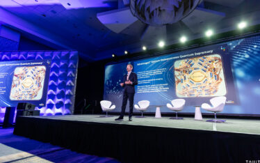 A man in a suit stands on stage near three white chairs, presenting slides about quantum supremacy to an audience in a large, modern conference room with blue lighting—captured by an expert Corporate Event Photographer Phoenix.