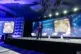 A man in a suit stands on stage near three white chairs, presenting slides about quantum supremacy to an audience in a large, modern conference room with blue lighting—captured by an expert Corporate Event Photographer Phoenix.