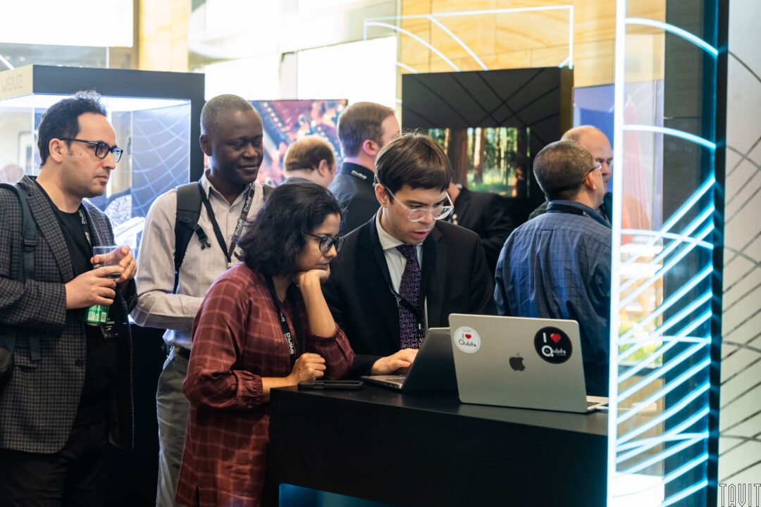 A group of people stands around a high table, focusing on a laptop covered with stickers. One person types while others watch intently. The setting appears to be a tech or business event with modern displays.