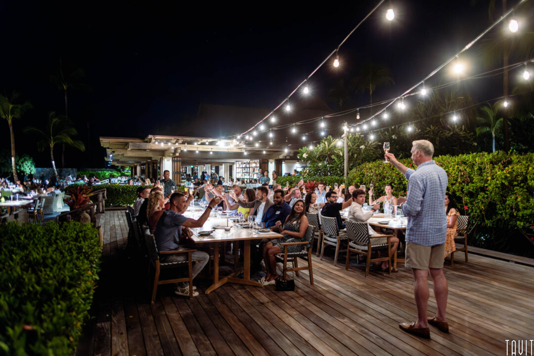 A group enjoys an evening under string lights as a man toasts them on a wooden deck surrounded by lush greenery—an ideal scene for event photography by a skilled corporate event photographer.