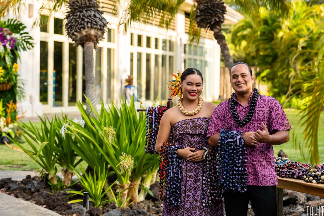 A smiling man and woman in patterned purple clothing stand outdoors with necklaces draped over their arms, surrounded by lush greenery and palm trees, captured by a skilled Corporate Event Photographer Phoenix.