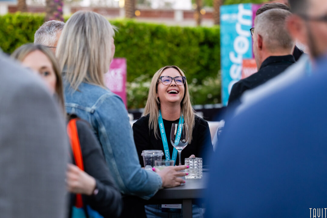 A woman with glasses and a conference lanyard laughs while sitting at a table outdoors, surrounded by others. The background features greenery and colorful event signs, capturing the lively atmosphere with expert Event Photography Phoenix.