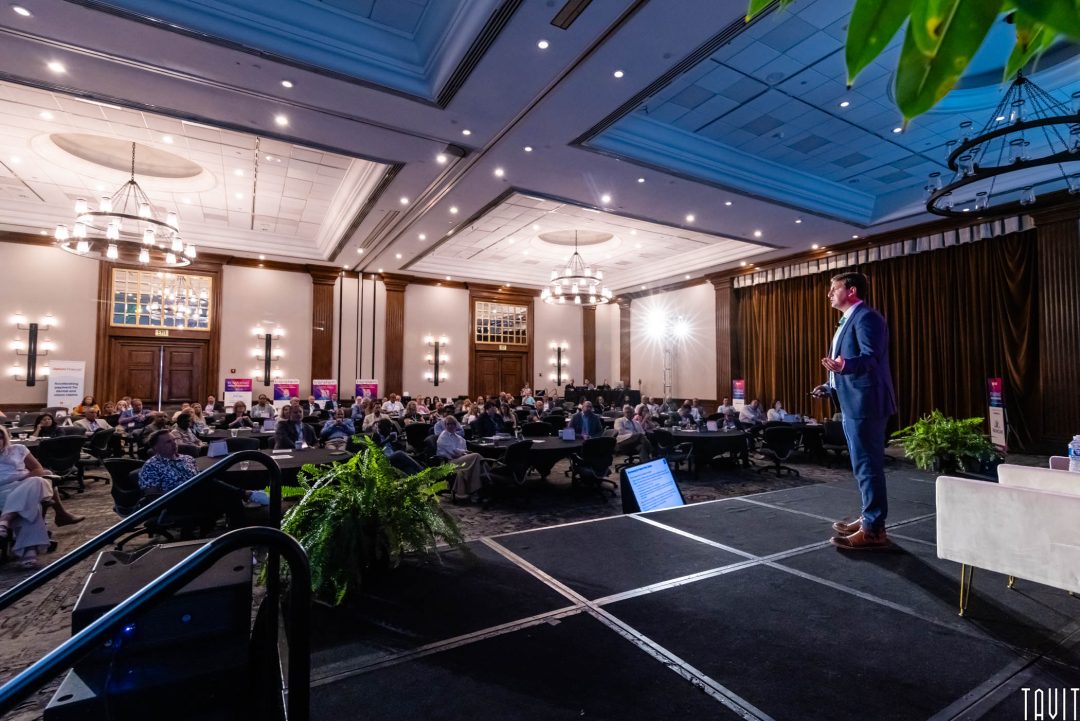 A Corporate Event Photographer captures a man in a suit speaking to a large seated audience in a spacious, well-lit conference room adorned with chandeliers and decorative plants.