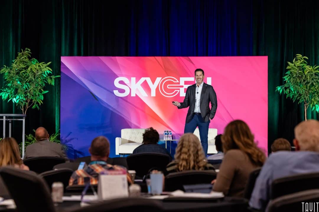 A man in a suit speaks on stage in front of a colorful "SKYGEN" backdrop, addressing an audience at a conference. Two white chairs and green plants enhance the scene—an ideal moment for an Event Photography Phoenix professional to capture.