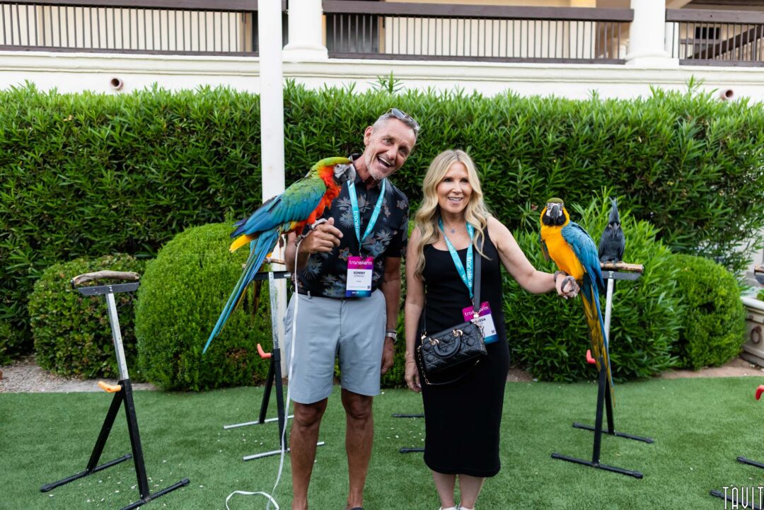 A man and woman smiling outdoors, each holding a colorful parrot. Standing on grass with greenery and building columns behind them, they wear event badges—captured by a Corporate Event Photographer in Phoenix.