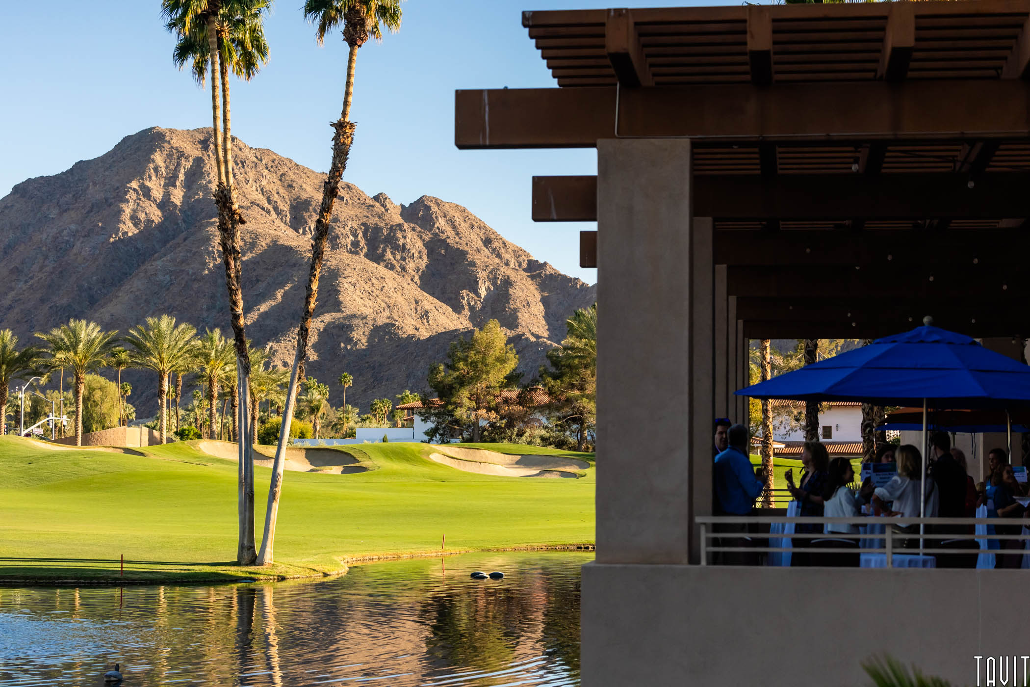 Professional outdoor corporate networking event at a luxury desert resort in Palm Springs featuring a modern patio, high tables, and palm trees against a dramatic mountain backdrop at sunset.
