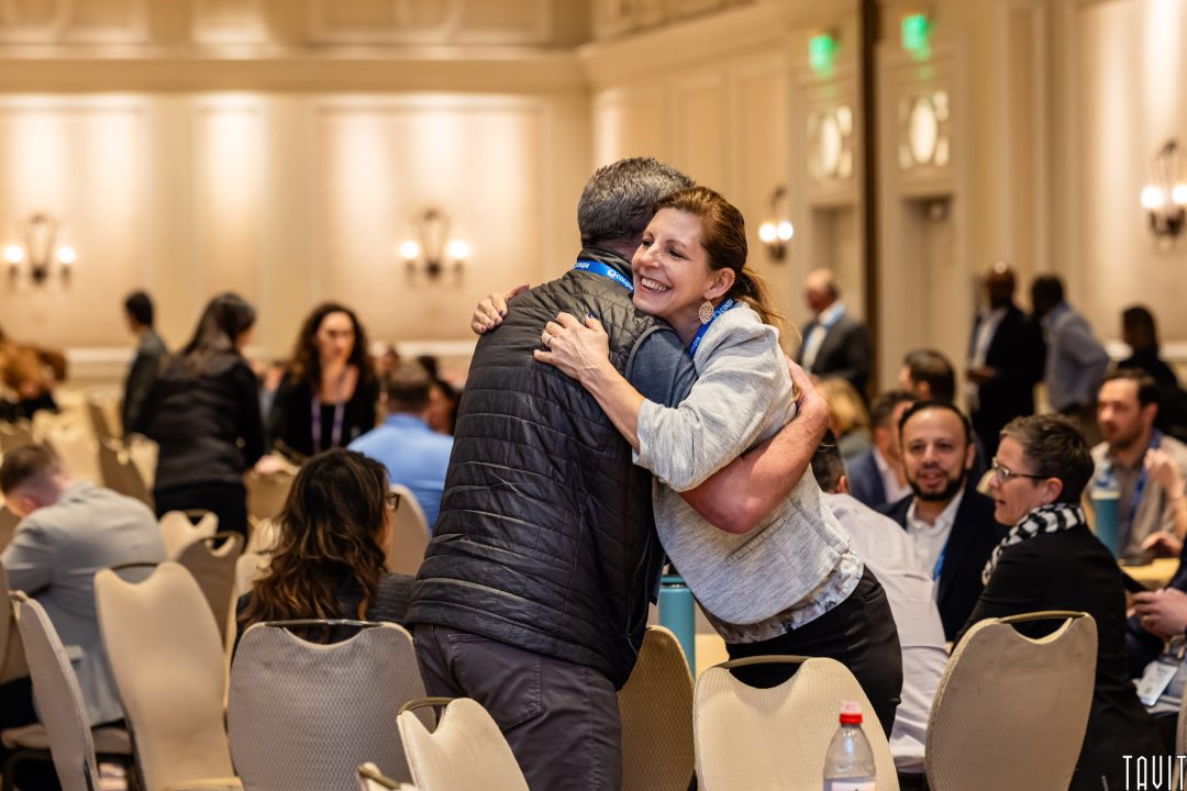 A woman and man hug and smile at a conference event, surrounded by other attendees seated and chatting in a large, well-lit room.