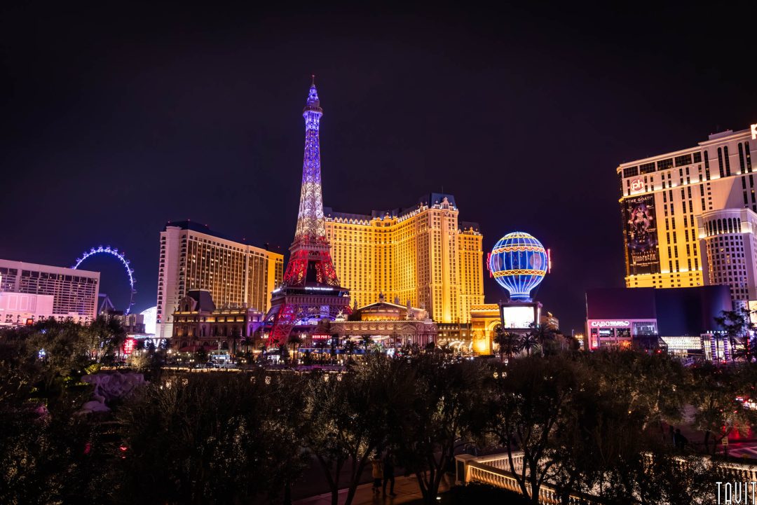 Las Vegas Strip at night featuring the illuminated replica Eiffel Tower, bright hotels, and colorful neon lights against a dark sky. Trees are visible in the foreground.