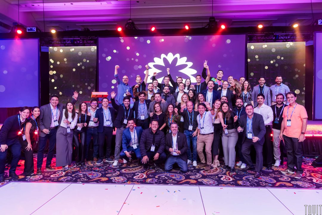A large group of people in business attire pose and smile together on a decorated stage with a purple backdrop and flower graphic, suggesting a celebratory or award event.