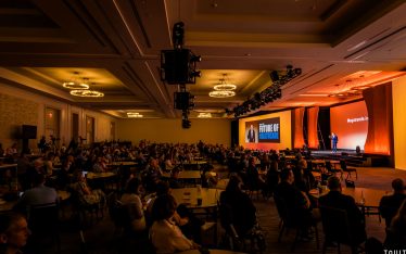 A large audience sits at round tables in a dimly lit conference room, watching a speaker on stage with a screen displaying "The Future of Healthcare." Warm lights illuminate the stage area.