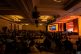 A large audience sits at round tables in a dimly lit conference room, watching a speaker on stage with a screen displaying "The Future of Healthcare." Warm lights illuminate the stage area.