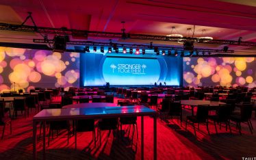 Large conference room with rows of chairs and tables facing a brightly lit stage. The backdrop reads "Stronger Together" with colorful circular light patterns projected across the walls, perfectly captured by a professional Scottsdale event photographer.