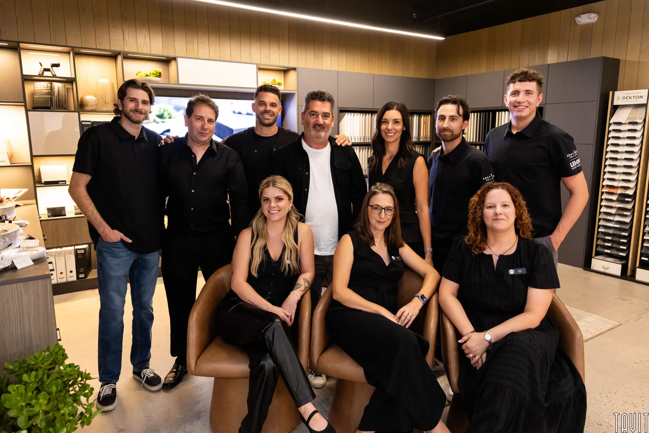 Eleven people pose together in a modern showroom with shelves and samples in the background; most are standing, while three are seated at the front, all smiling and casually dressed.