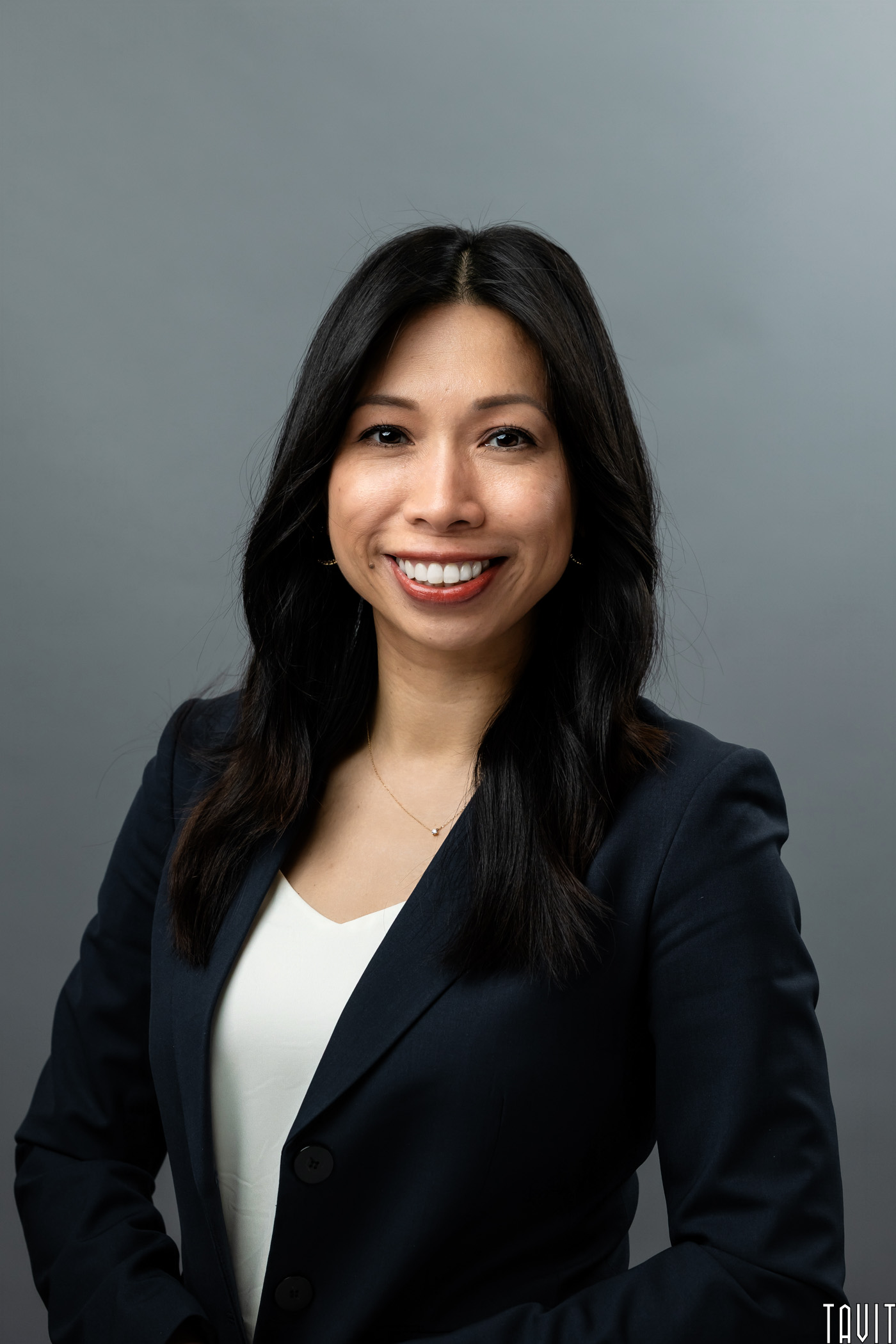 A woman with long dark hair, wearing a navy blazer and white top, smiles at the camera against a plain gray background.