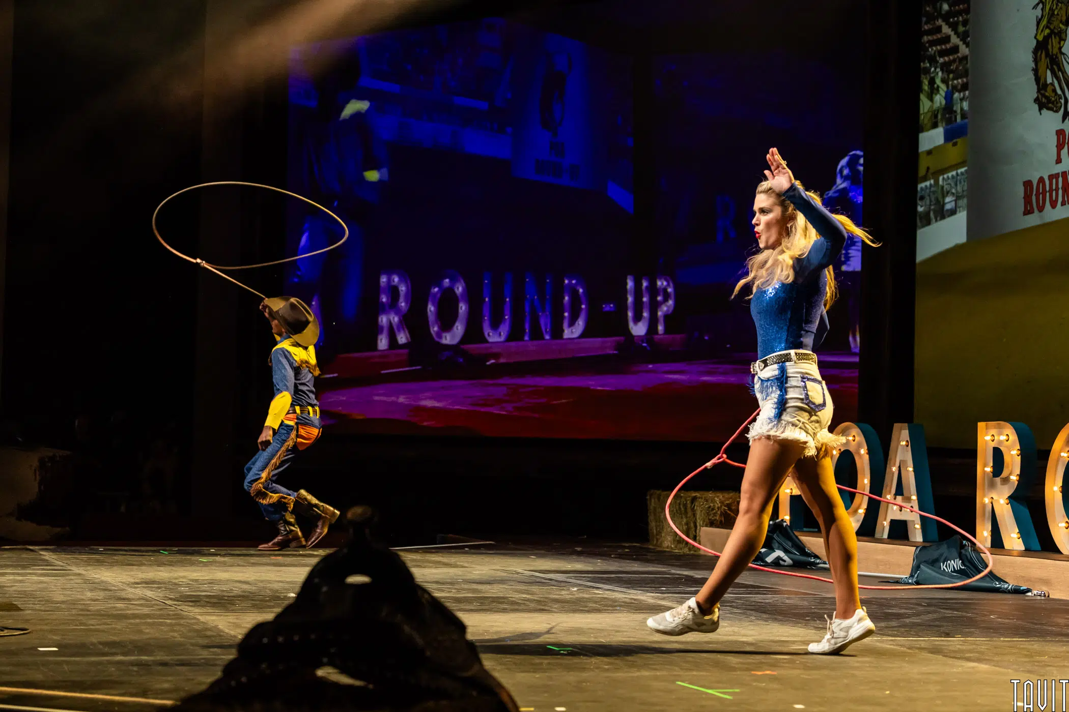 A woman in shorts and boots and a child in cowboy attire perform rope tricks on stage, with "ROUND-UP" letters and rodeo visuals visible in the background.