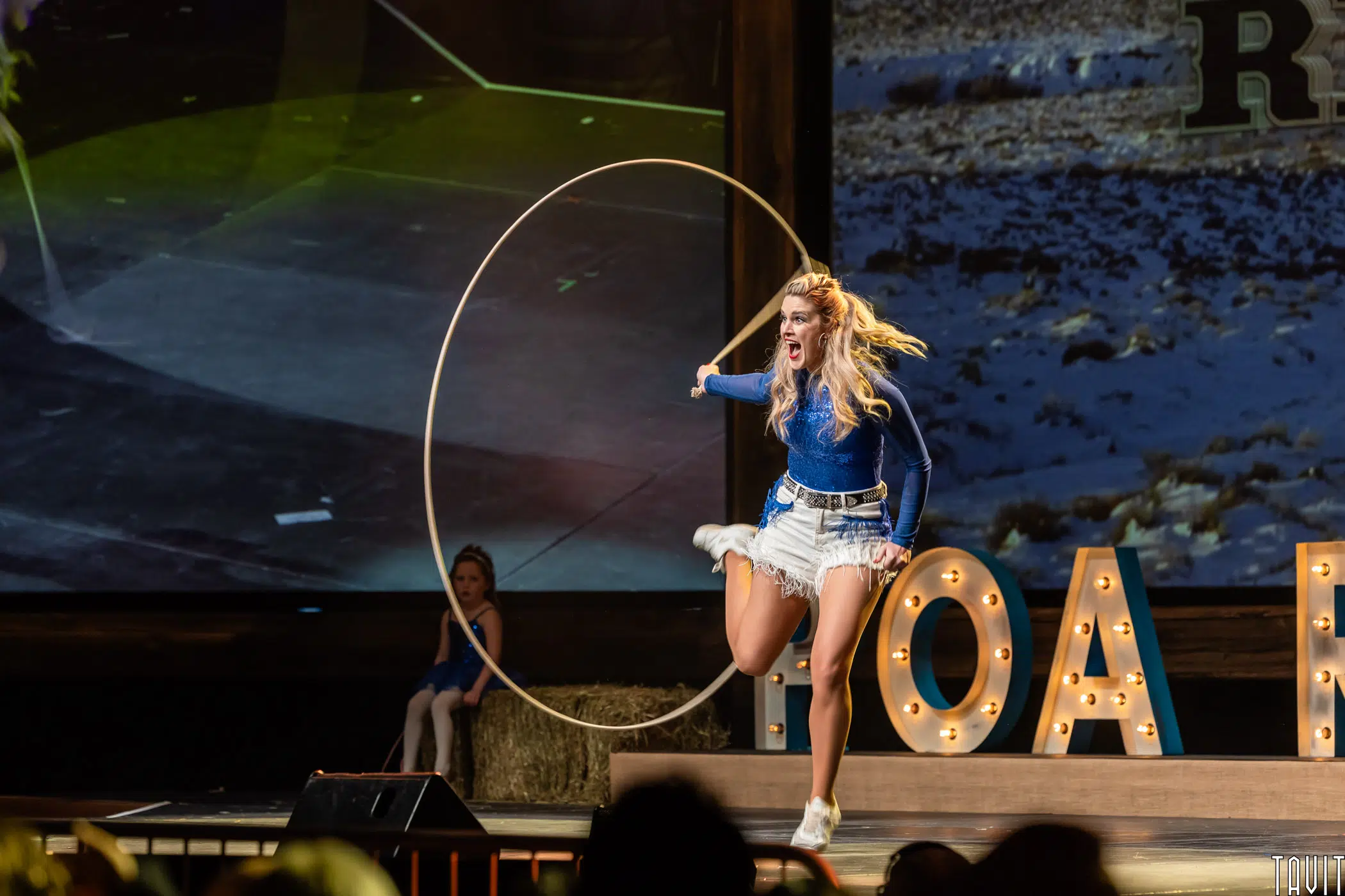 A woman in a blue shirt and white shorts jumps energetically through a large lasso on stage, with another girl seated in the background and large lit-up letters behind them.