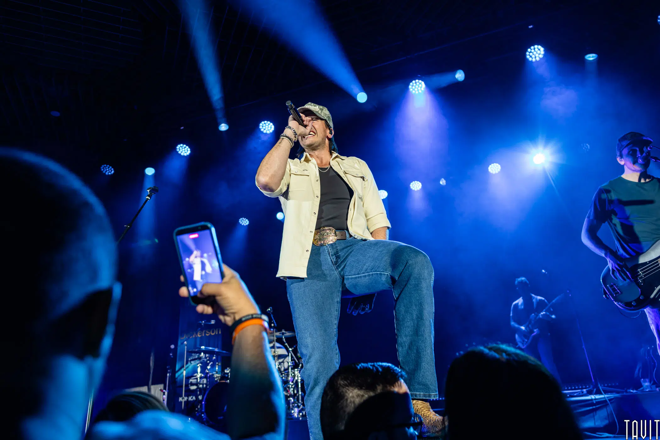 A singer in a beige shirt and cap performs on stage under blue lights, holding a microphone. An audience member in the foreground films with a phone. Band members and instruments are visible in the background.