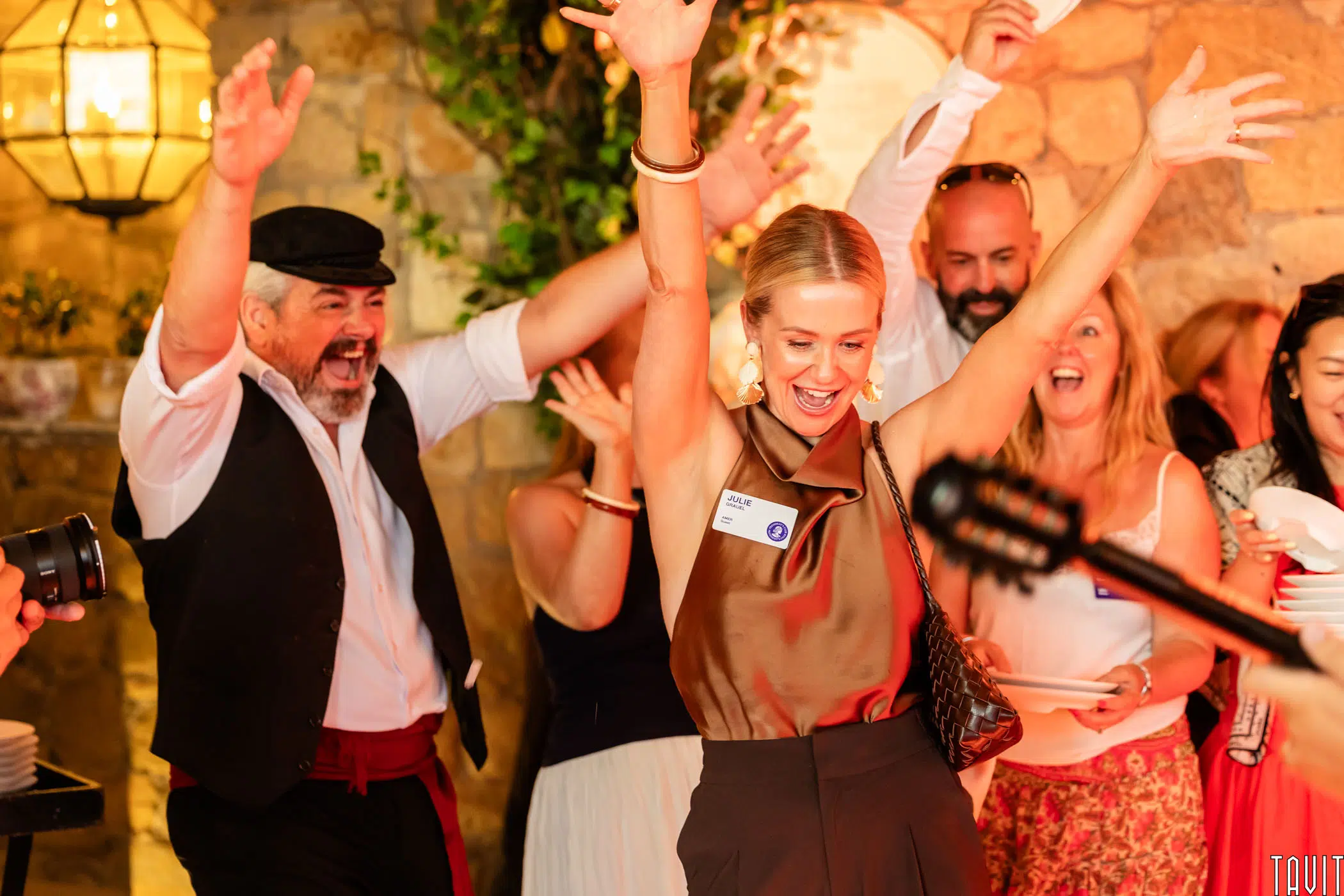 A group of people joyfully celebrate indoors. A woman in front raises her arms and smiles, surrounded by others cheering and clapping. Warm lighting and stone walls create a festive atmosphere.