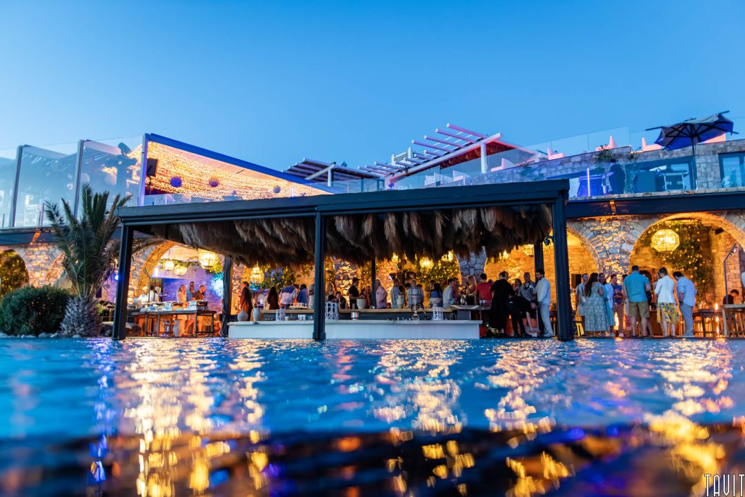 A lively outdoor bar and restaurant by a pool at sunset, with people socializing under string lights and reflections shimmering on the water.
