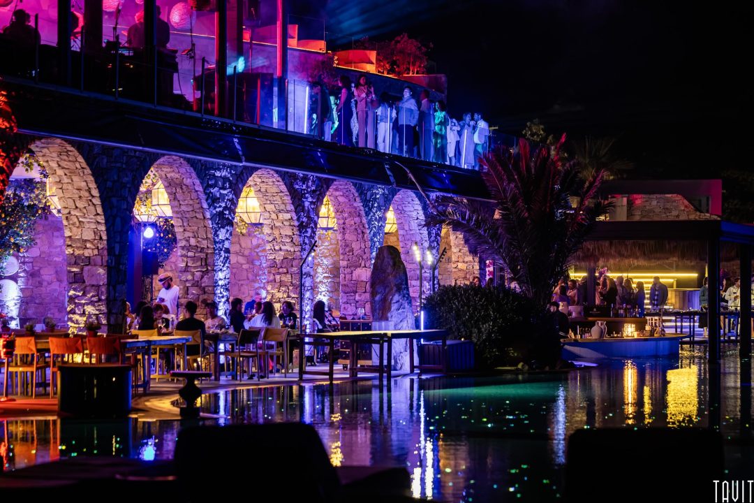 A vibrant nightlife scene at a poolside venue with stone arches, colorful lights, people dining, and guests on an upper balcony, all reflecting on the water at night.