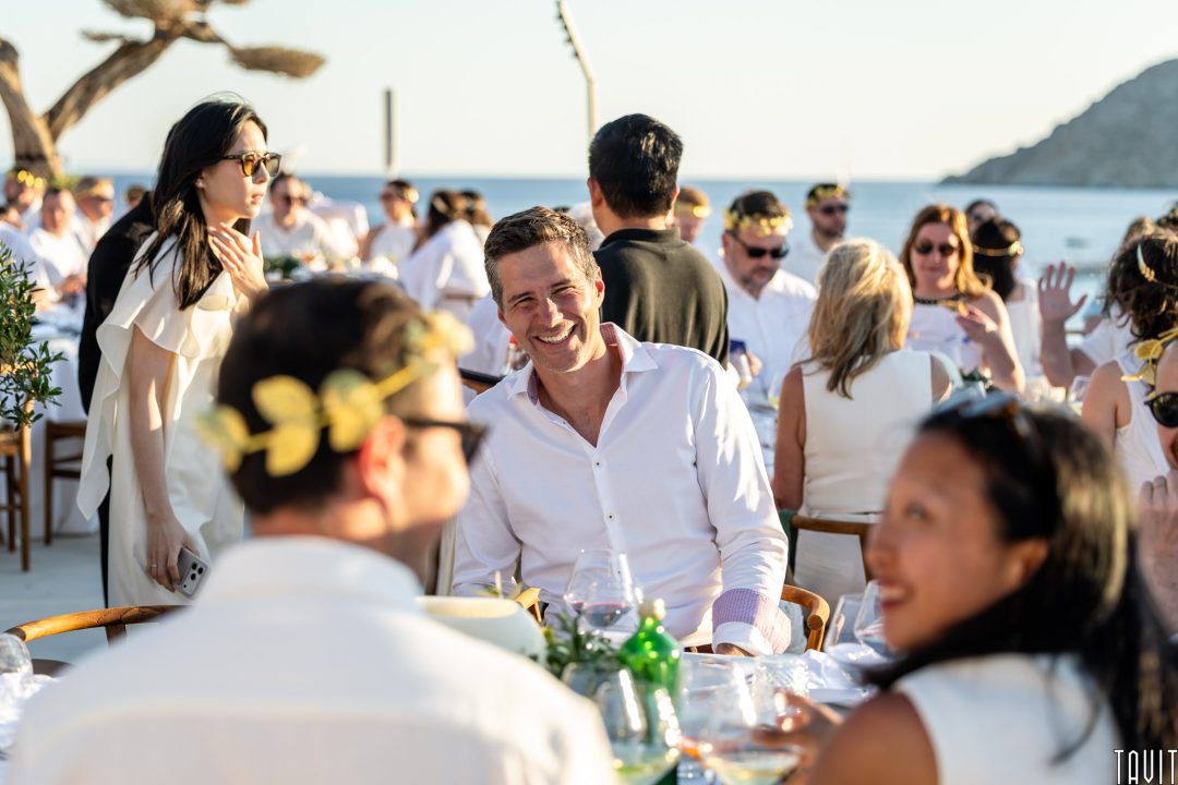 A man in a white shirt smiles at an outdoor seaside gathering, surrounded by people in white outfits and yellow laurel headbands, with sunlight and the sea in the background.