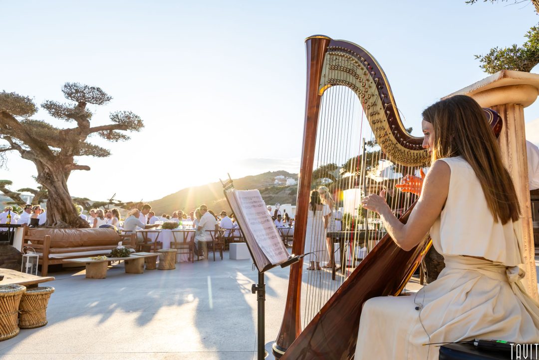 A woman in a cream dress plays a harp outdoors at sunset with guests seated at tables in the background, surrounded by trees and mountains.