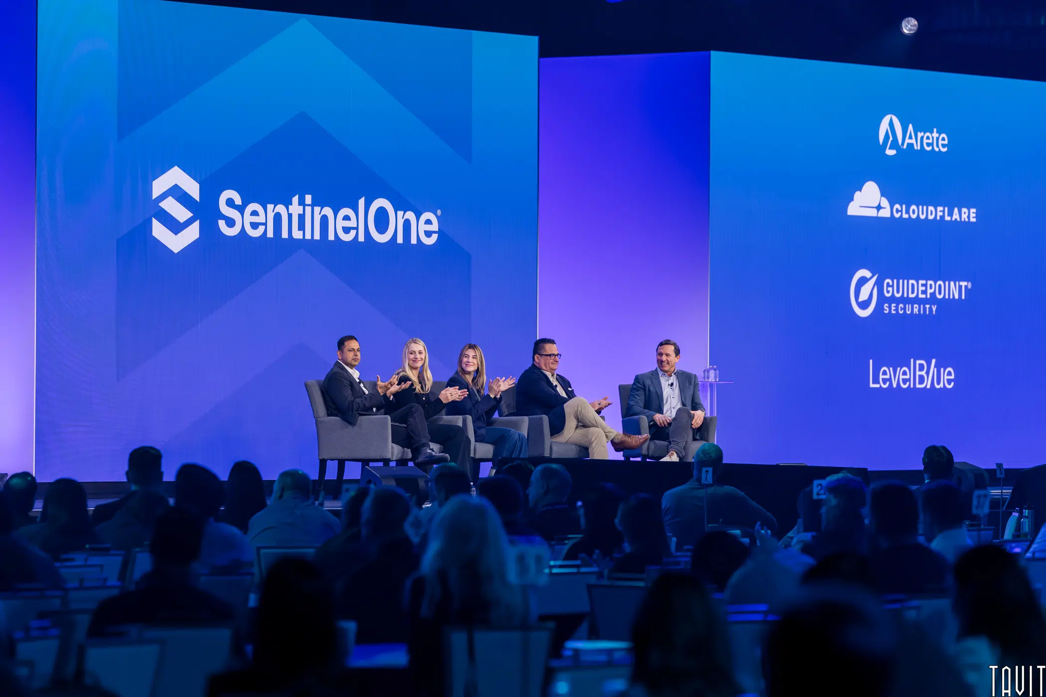 Four people sit on stage at a tech conference panel. Behind them, large blue screens display the SentinelOne logo and partner logos: Arete, Cloudflare, GuidePoint Security, and LevelBlue. Audience members watch.