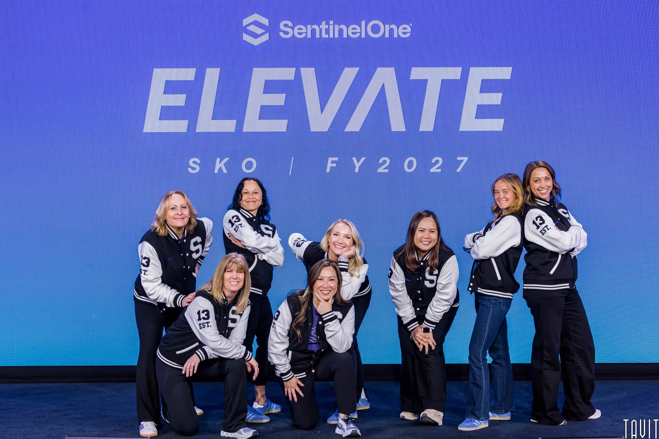 Eight women in black and white varsity jackets pose and smile in front of a blue backdrop that reads "SentinelOne ELEVATE SKO | FY2027.
