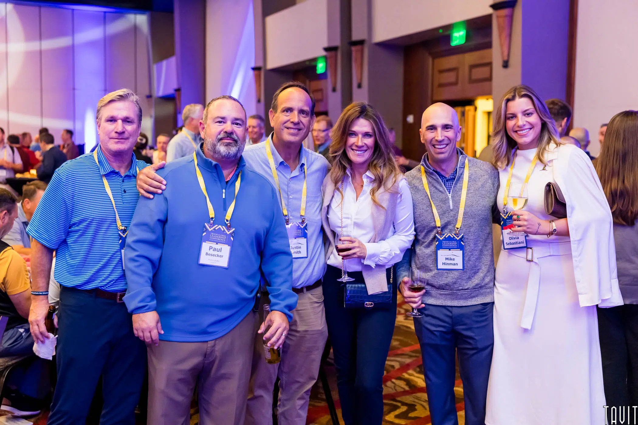 Six adults pose together at an indoor event, smiling at the camera. All wear event badges and business casual attire. The background shows other attendees and event decor.
