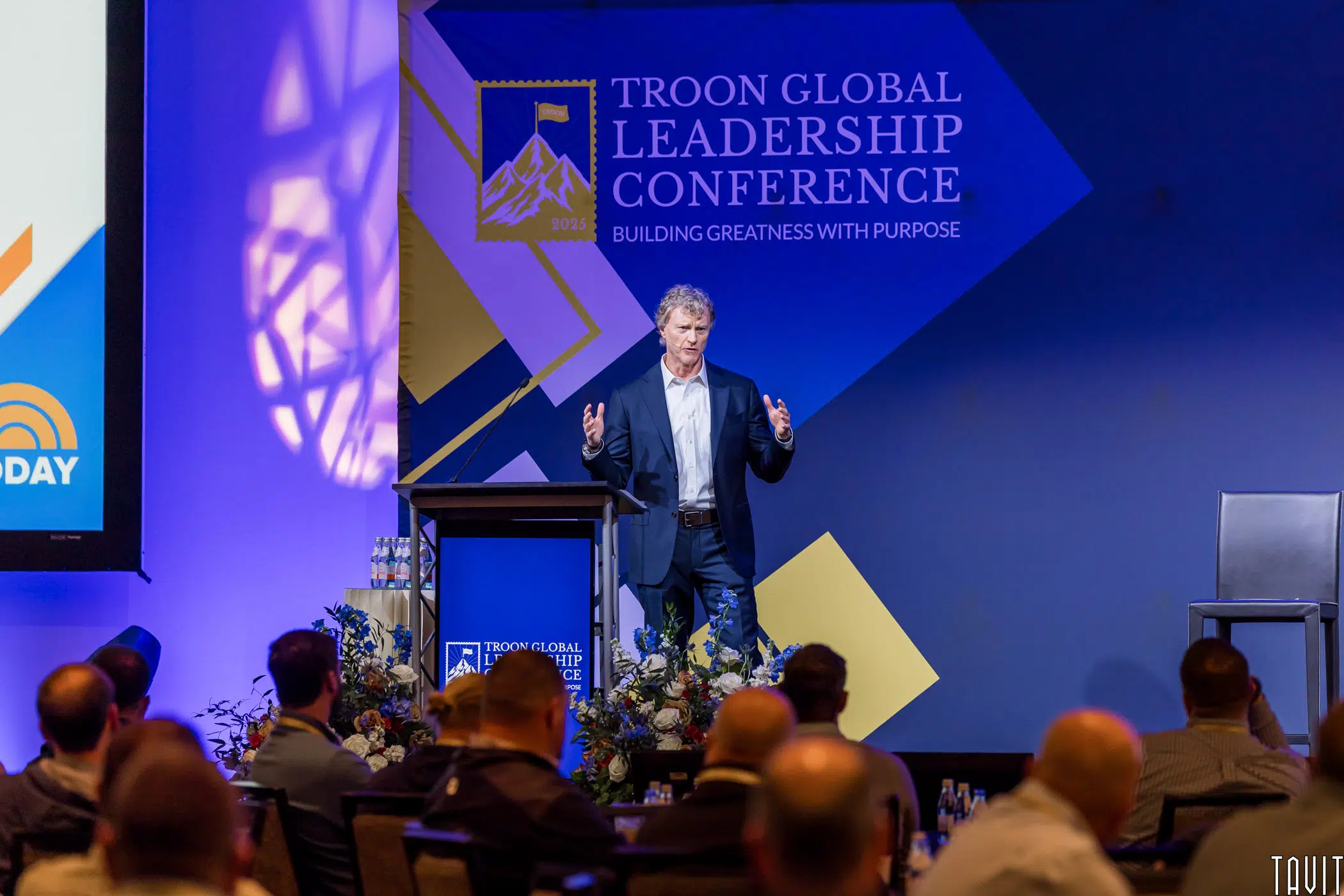 A man speaks on stage at the Troon Global Leadership Conference. Attendees watch from their seats, and the event banner and floral arrangements are visible in the background.