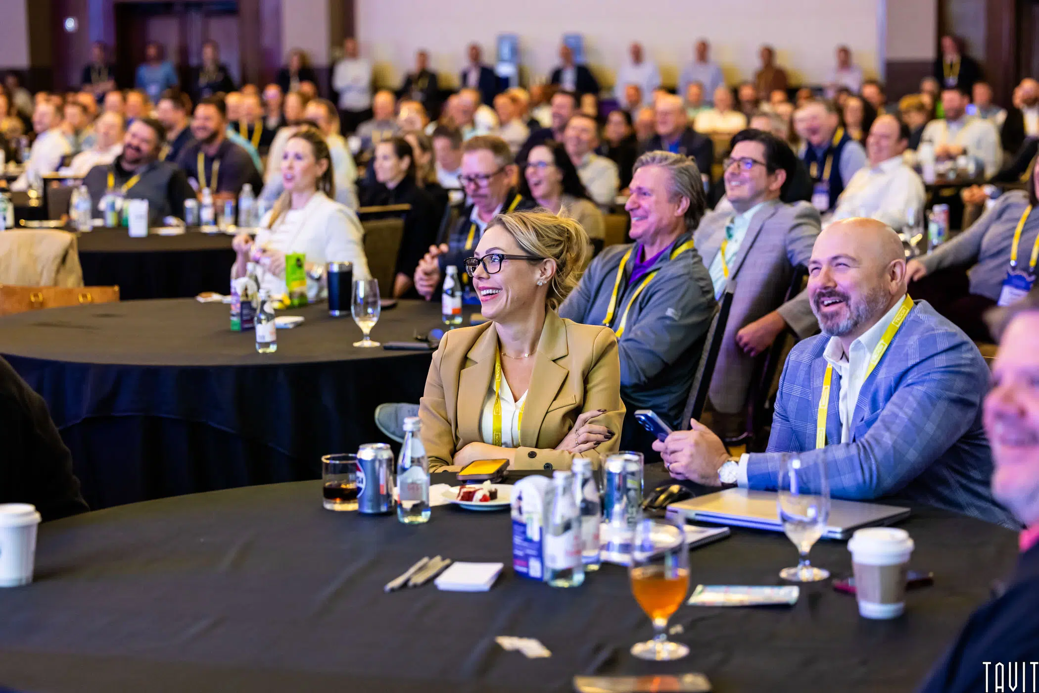A large group of people sits at round tables in a conference room, watching a presentation and smiling. Many wear conference badges and have drinks on the tables. The atmosphere appears lively and engaged.