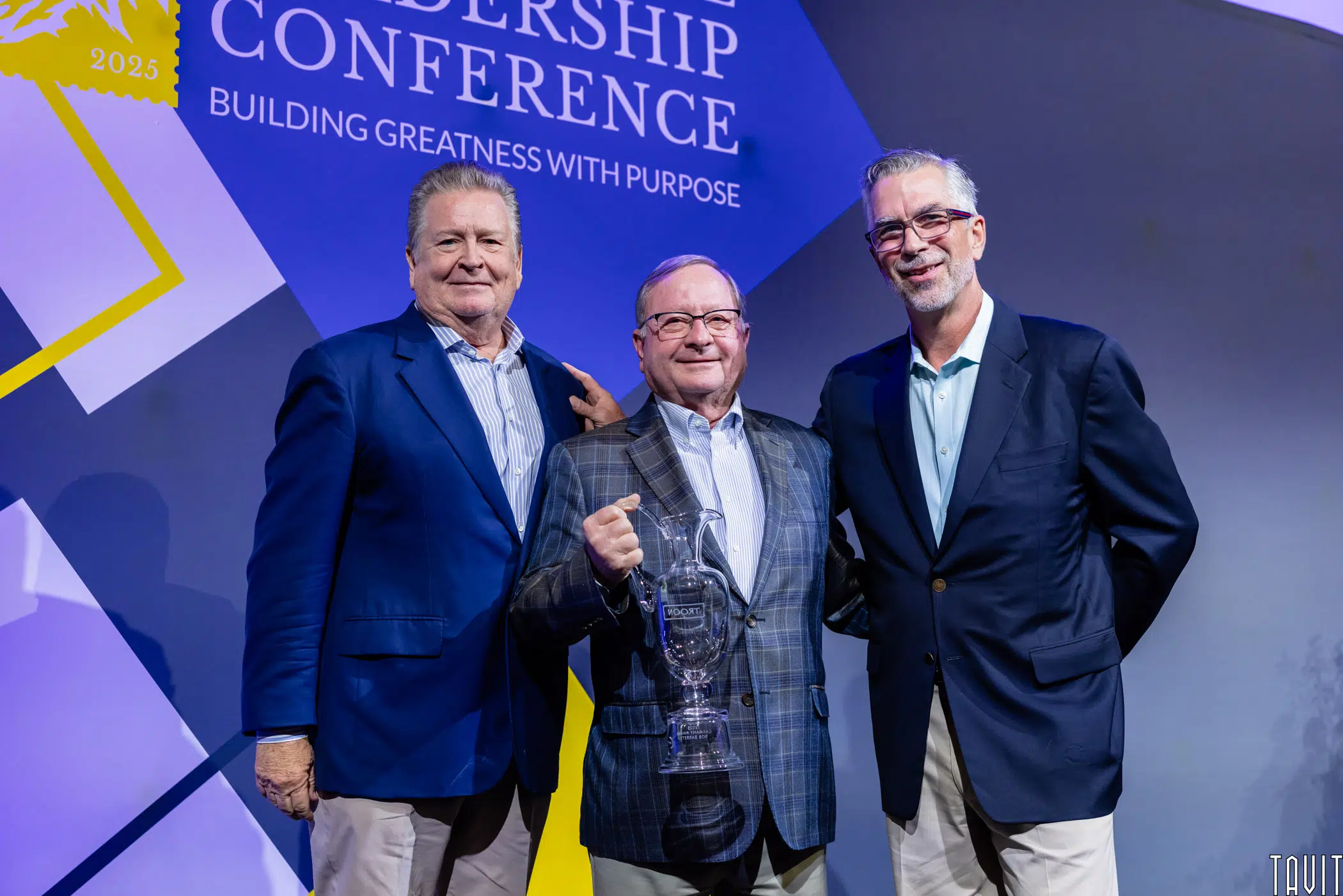 Three men in suits stand on stage at a leadership conference. The man in the center holds a glass trophy and smiles. A conference banner with the words “Building Greatness with Purpose” is visible in the background.