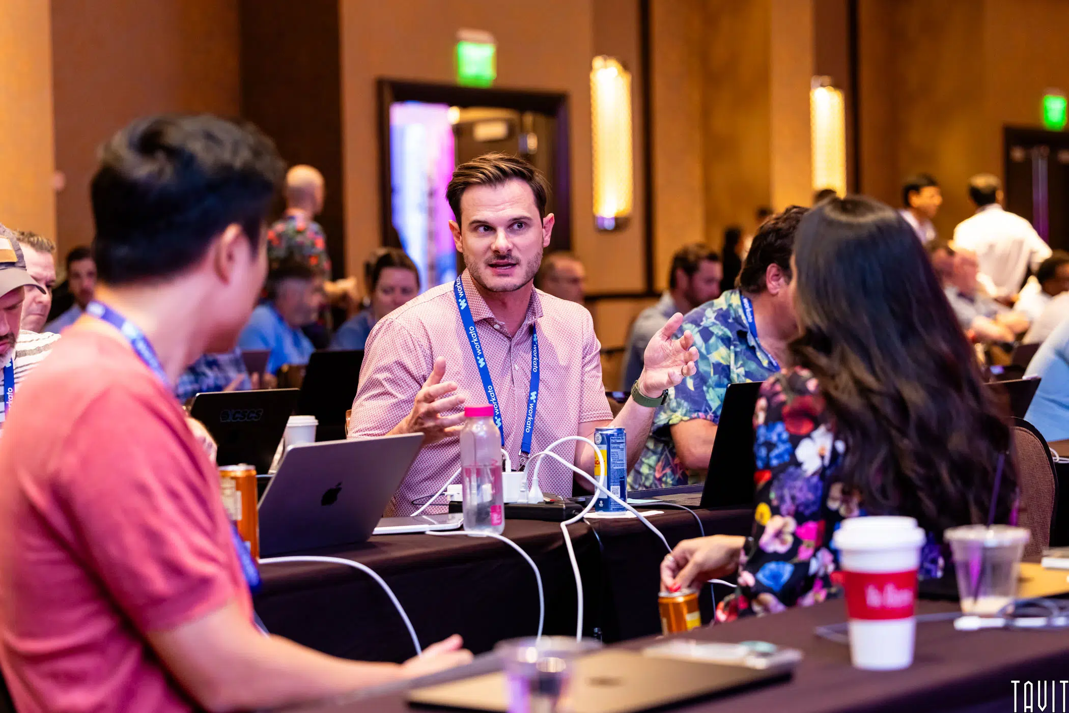 A group of people sit at a conference table with laptops, drinks, and notepads, engaged in discussion. One person gestures while speaking to others, creating an interactive and focused atmosphere.