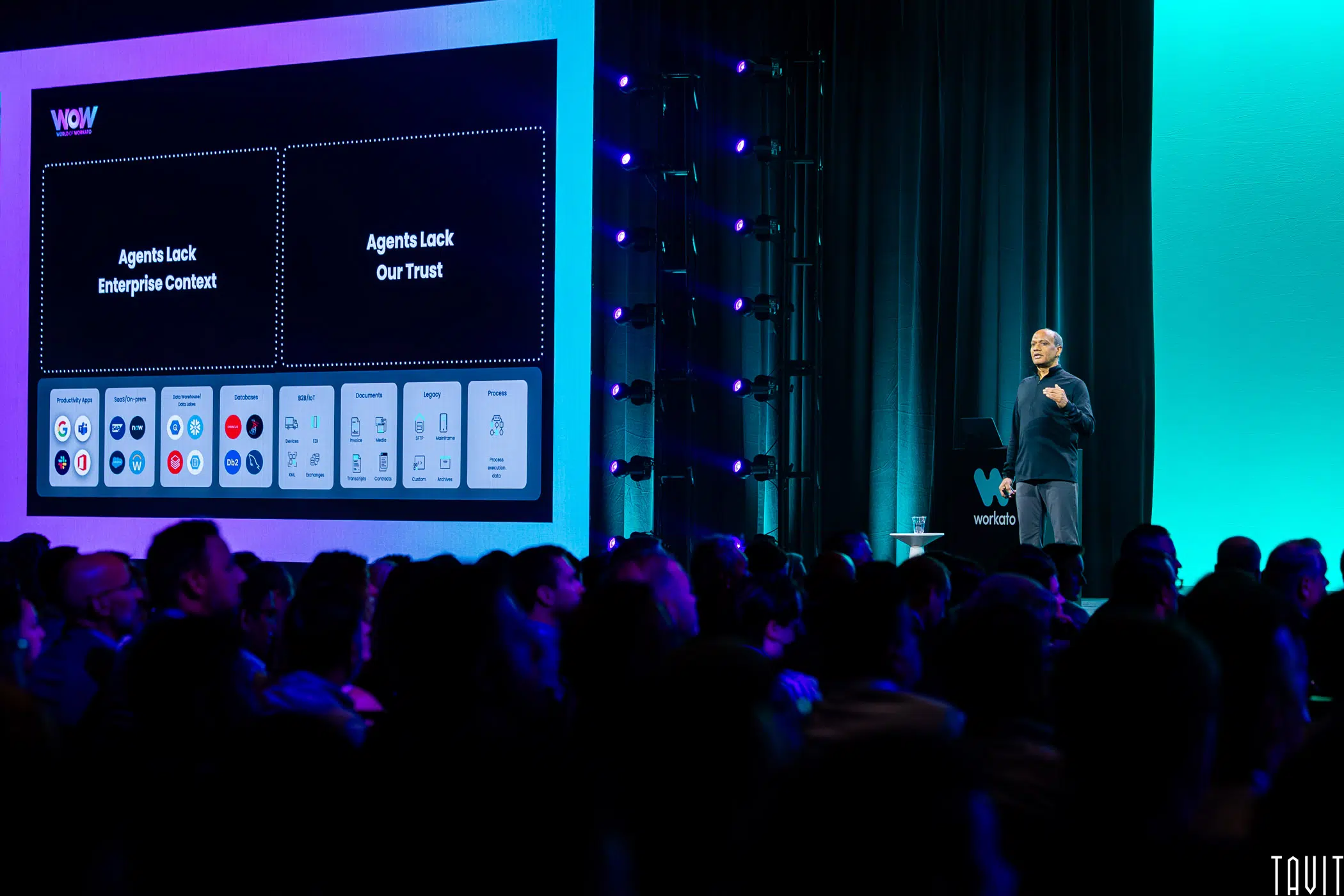 A speaker presents on stage to a seated audience, with a large screen displaying a tech-related slide titled "WOW" and two sections labeled "Agents Lack Enterprise Context" and "Agents Lack Our Trust.