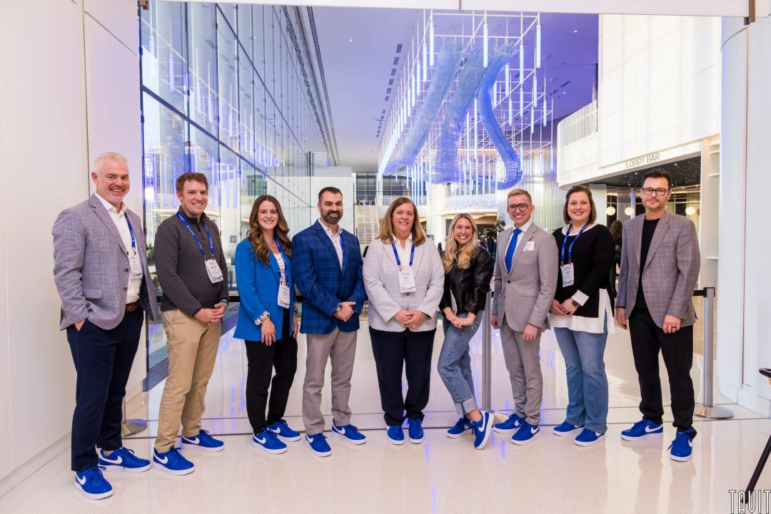 Ten people stand in a modern, brightly lit lobby, all dressed in business attire with blue sneakers, smiling at the camera under a large art installation with neon blue lights.