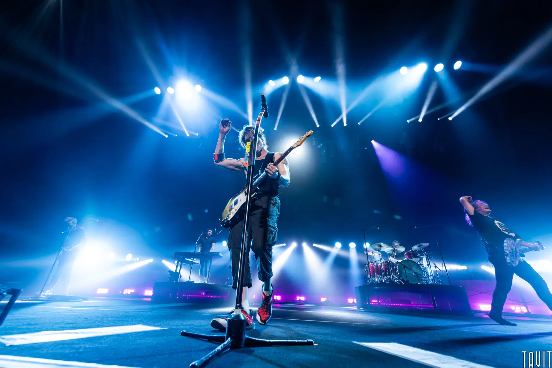 A rock band performs energetically on stage under bright blue and white lights, with one musician playing guitar at the front and others on drums, keyboard, and guitar in the background.