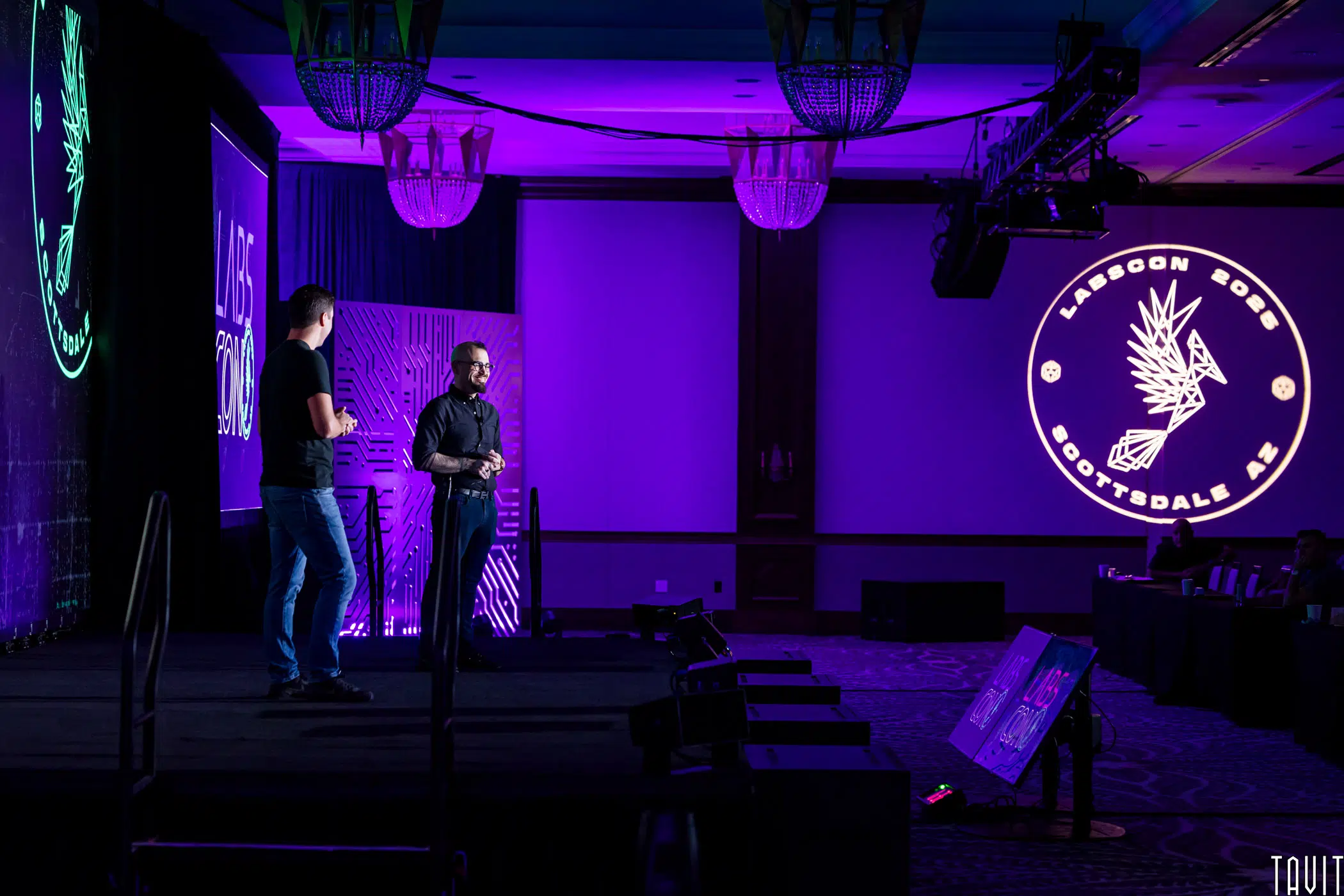 Two people stand and speak on a stage at a conference. The room is lit with purple lighting, and a large circular logo with a stylized eagle and the words “LABSCON 2022 SCOTTSDALE AZ” is projected on the wall.