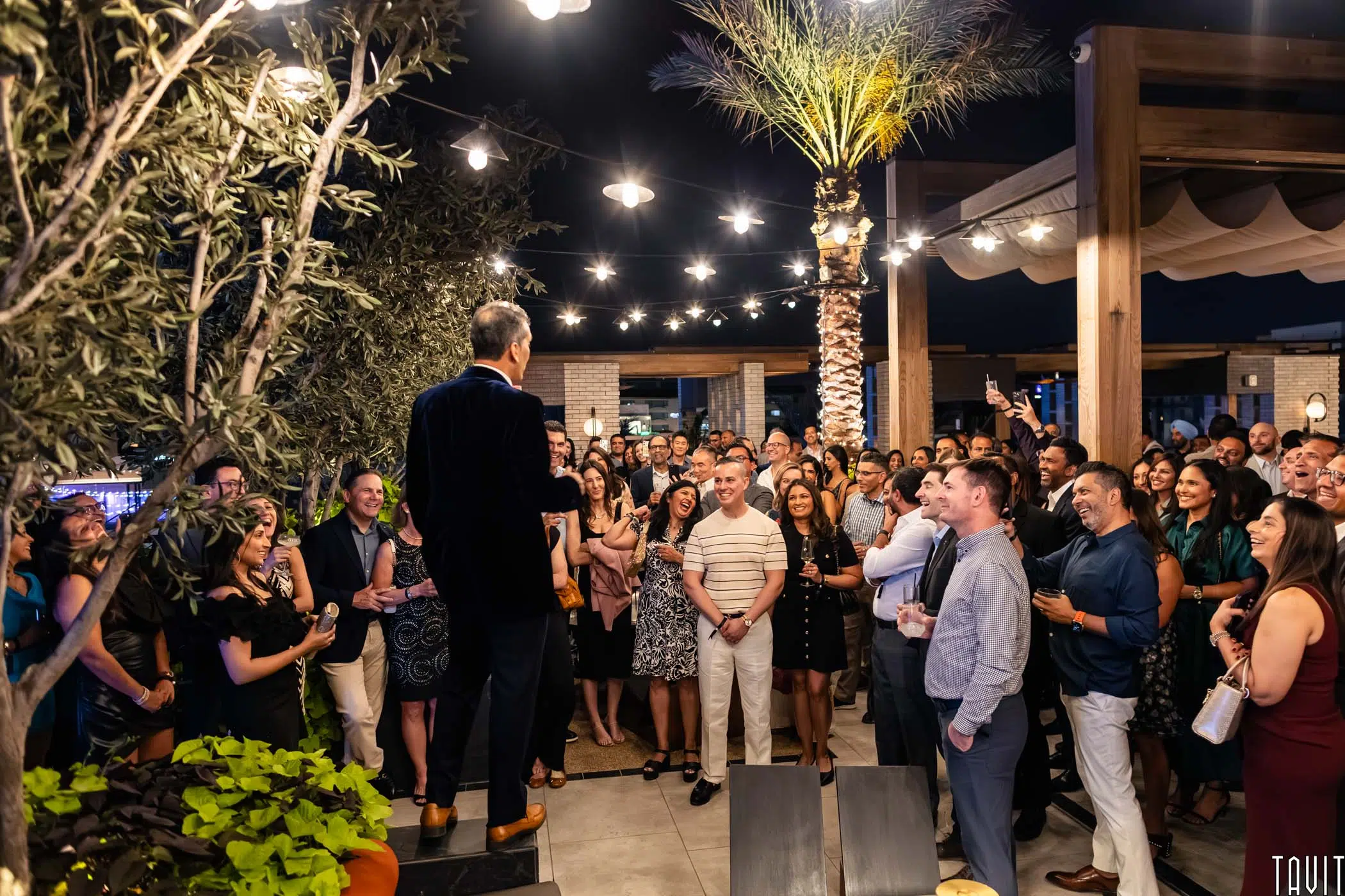 A man stands on a platform speaking to a large, smiling crowd at an outdoor evening event with palm trees, string lights, and modern decor.