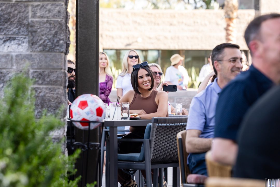 A woman with dark hair and sunglasses on her head smiles while sitting at a table outdoors. People around her are also enjoying themselves. A soccer ball rests nearby. The setting appears to be a casual gathering or event.