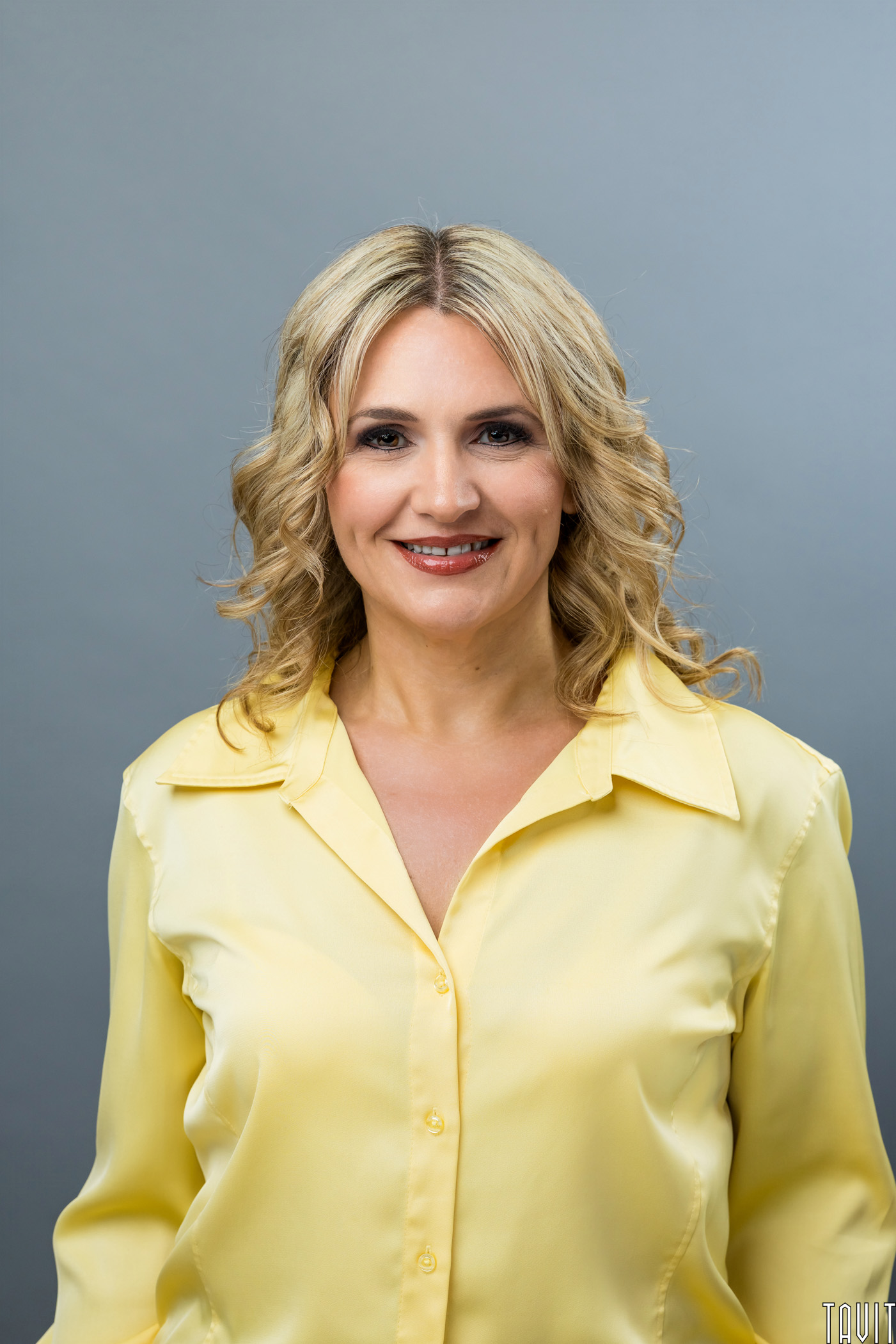Smiling woman with wavy blonde hair wearing a yellow button-up blouse, standing in front of a plain gray background.