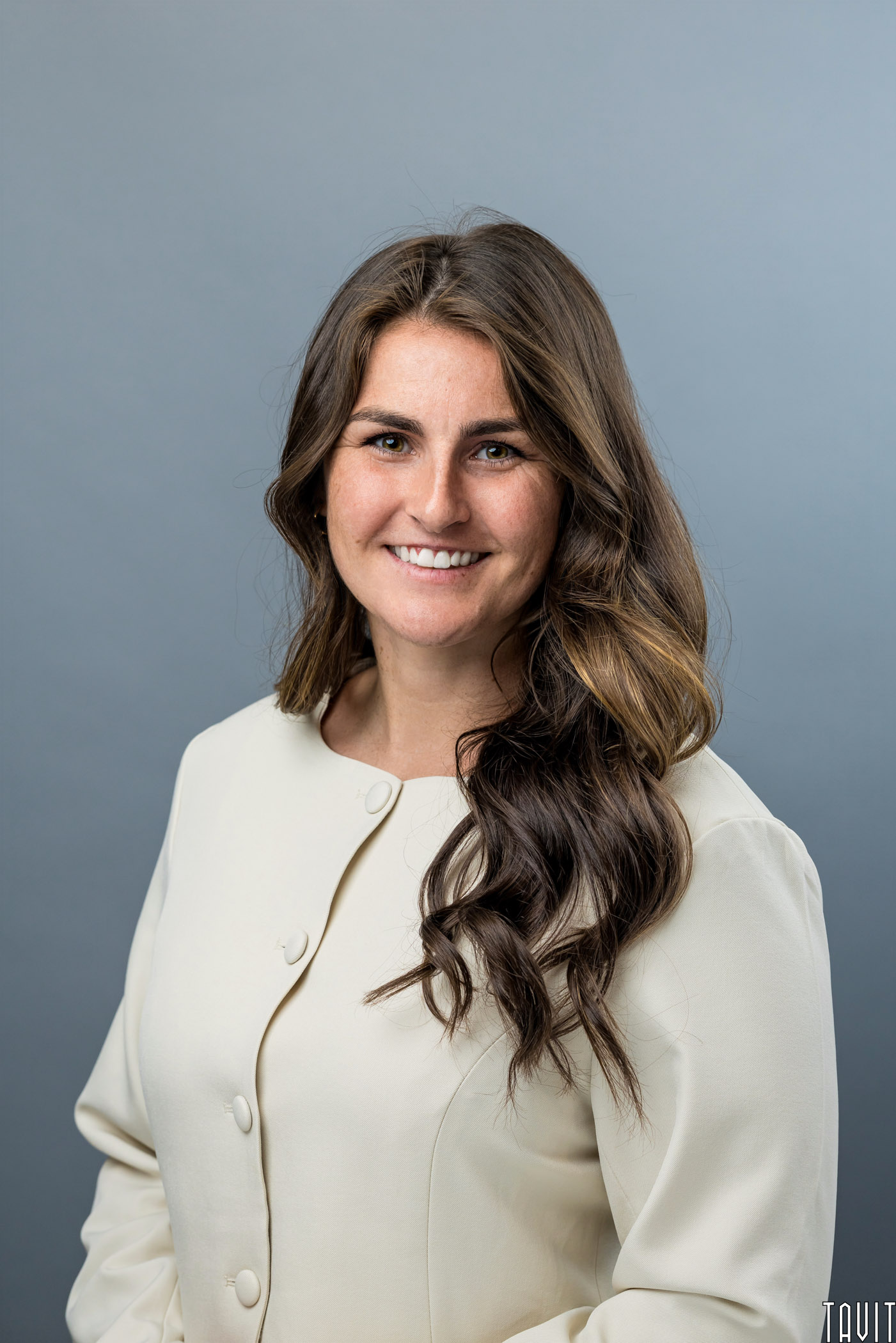 A woman with long brown wavy hair wearing a cream-colored blazer is smiling at the camera against a plain gray background.