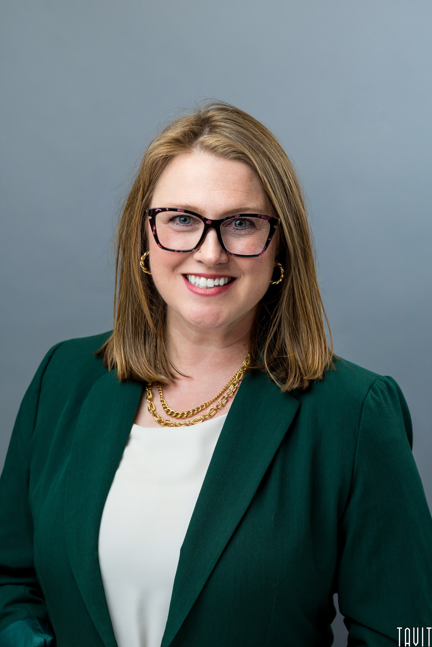 A woman with shoulder-length light brown hair, glasses, and a green blazer over a white shirt smiles against a plain gray background.
