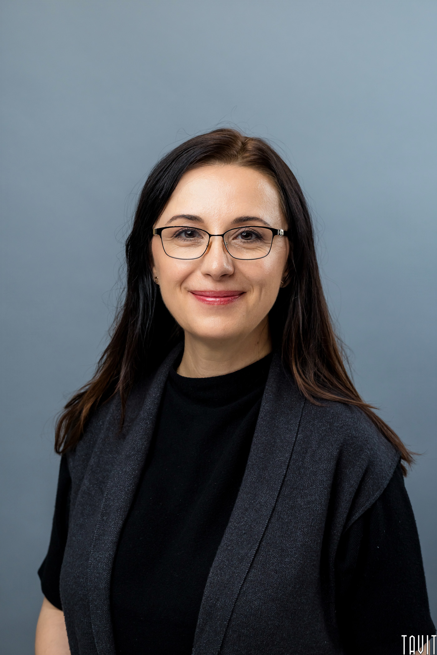 A woman with long dark hair and glasses smiles at the camera. She is wearing a black top and a dark gray vest, standing against a plain gray background.
