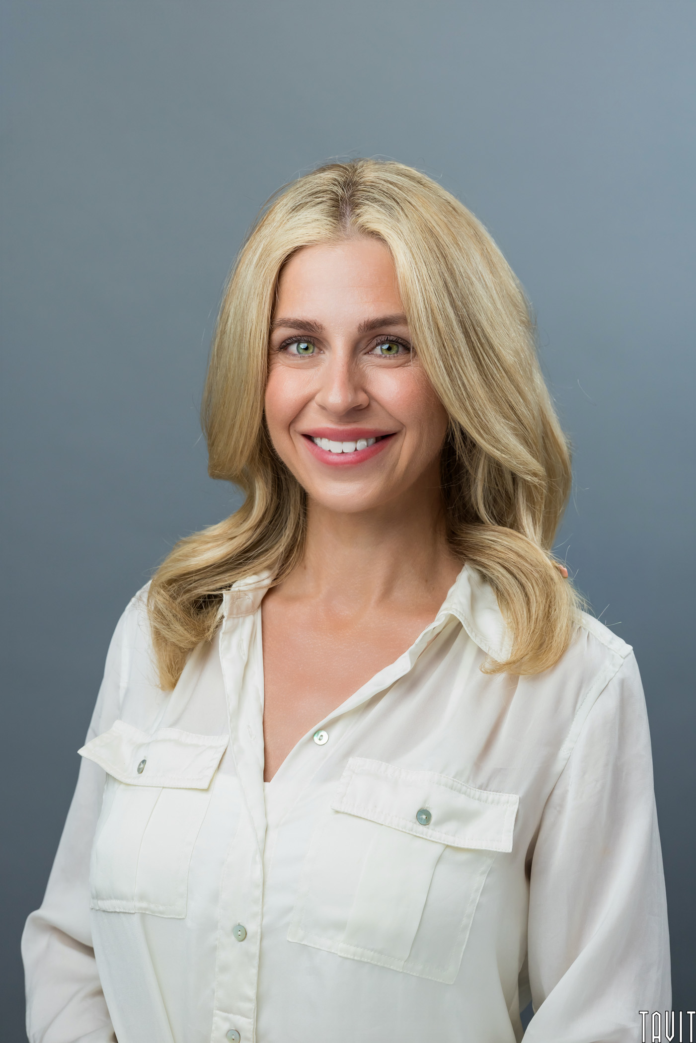 A woman with long blonde hair and blue eyes smiles at the camera while wearing a white button-up shirt, standing against a plain gray background.