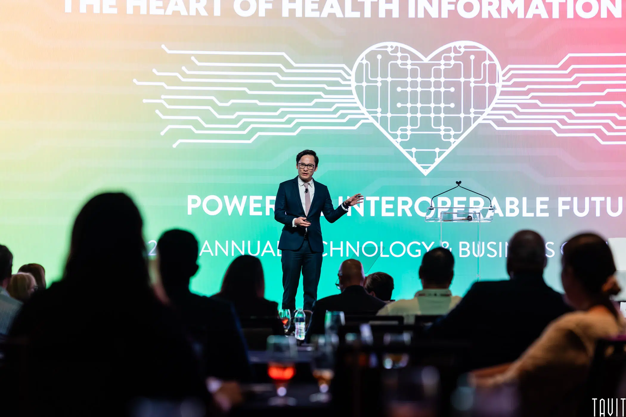 A speaker in a suit stands on stage in front of an audience, presenting with a large screen behind him displaying a digital heart graphic and the words "The Heart of Health Information.