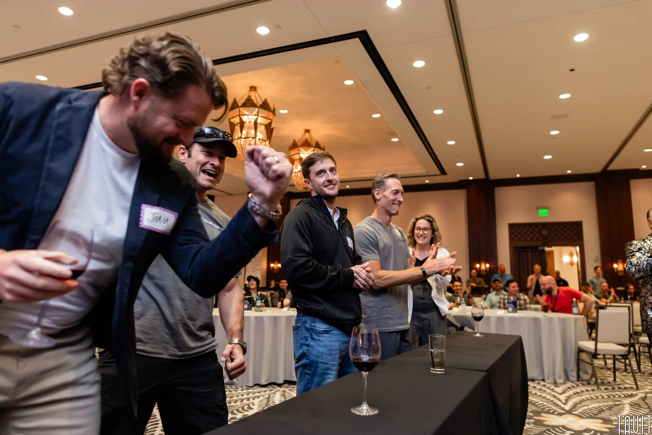 Six people stand and cheer at an indoor event, some clapping, one raising his fist in excitement. Glasses of wine are on the table in front of them, and attendees sit at round tables in the background.