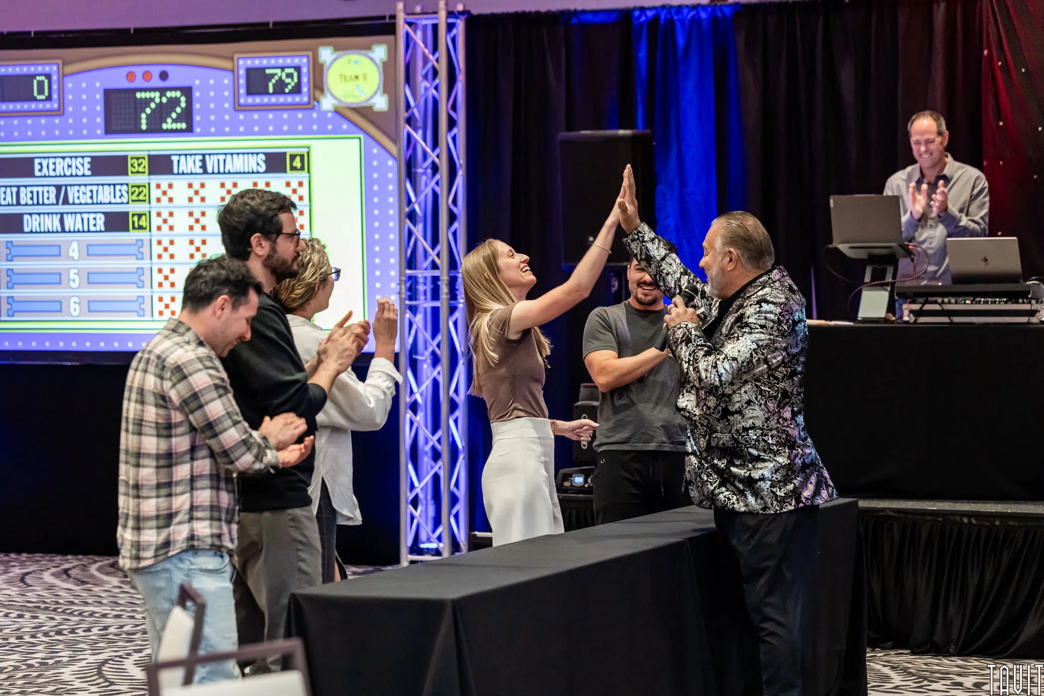 A group of people celebrate onstage at a game show event, with a host in a shiny jacket giving a high-five to a contestant while others clap and smile. A DJ stands at a booth in the background.