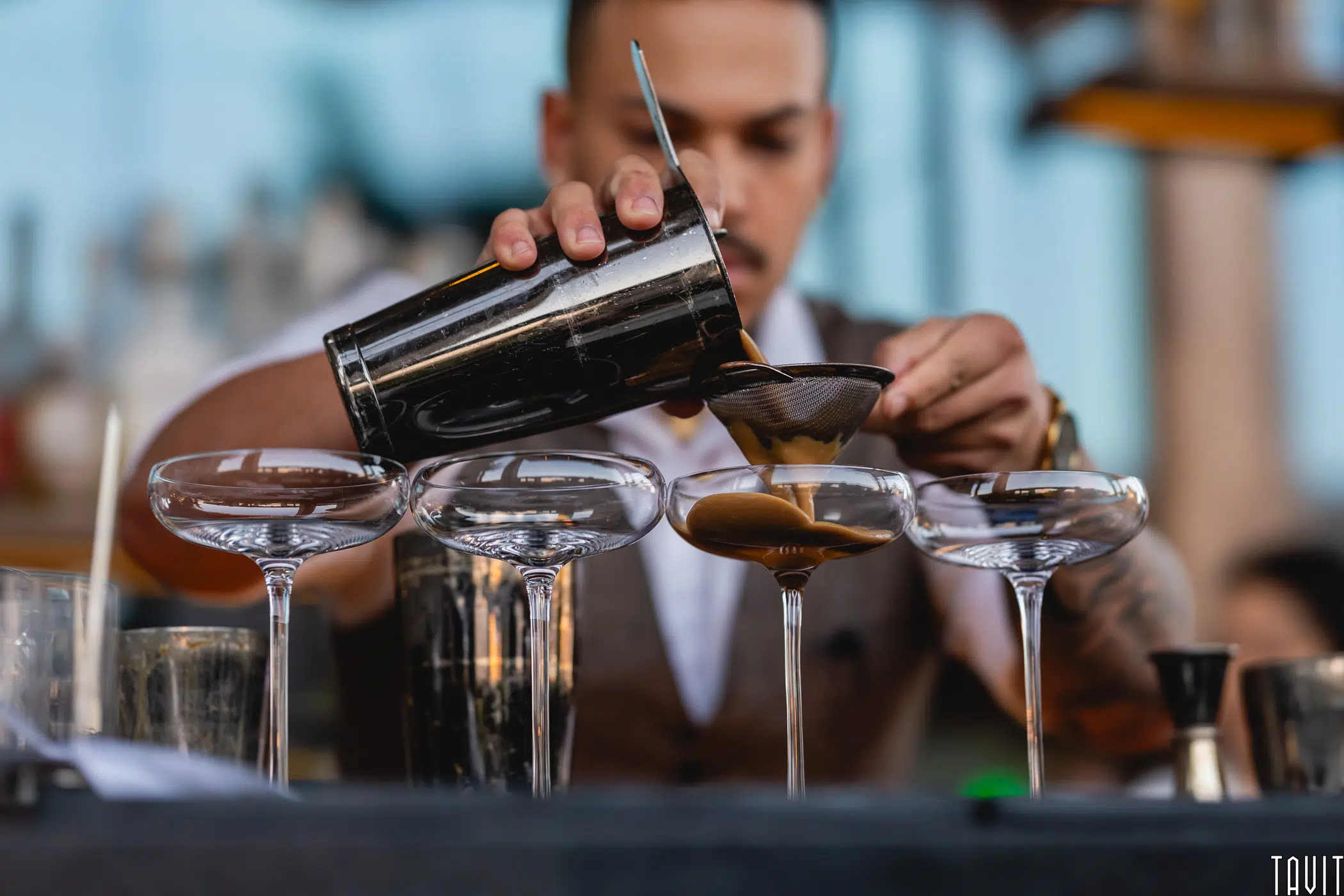 A bartender pours a brown cocktail through a strainer into one of three coupe glasses, focusing on precise presentation at a bar.